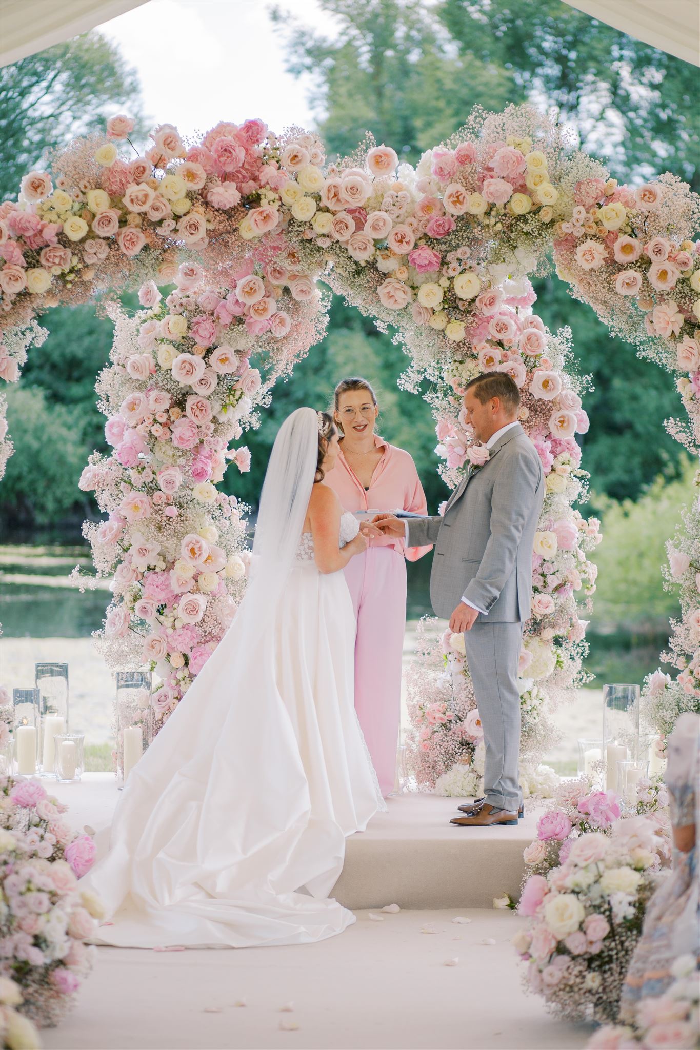 Celebrant Olivia Coleman stands alongside a bride and groom exchanging heartfelt vows by a beautiful arch of flowers at a wedding in Essex. The couple holds hands, surrounded by the enchanting atmosphere of love and commitment on their special day.